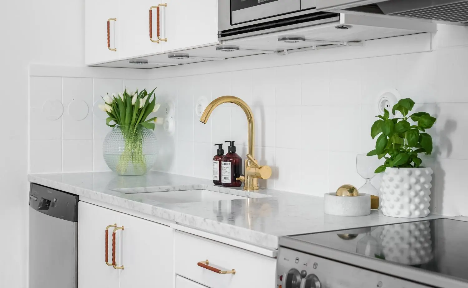 En köksbänk i vit marmor med en guldkran, en bukett vita tulpaner, en krukväxt och vita skåp med guldhandtag från en köksrenovering.